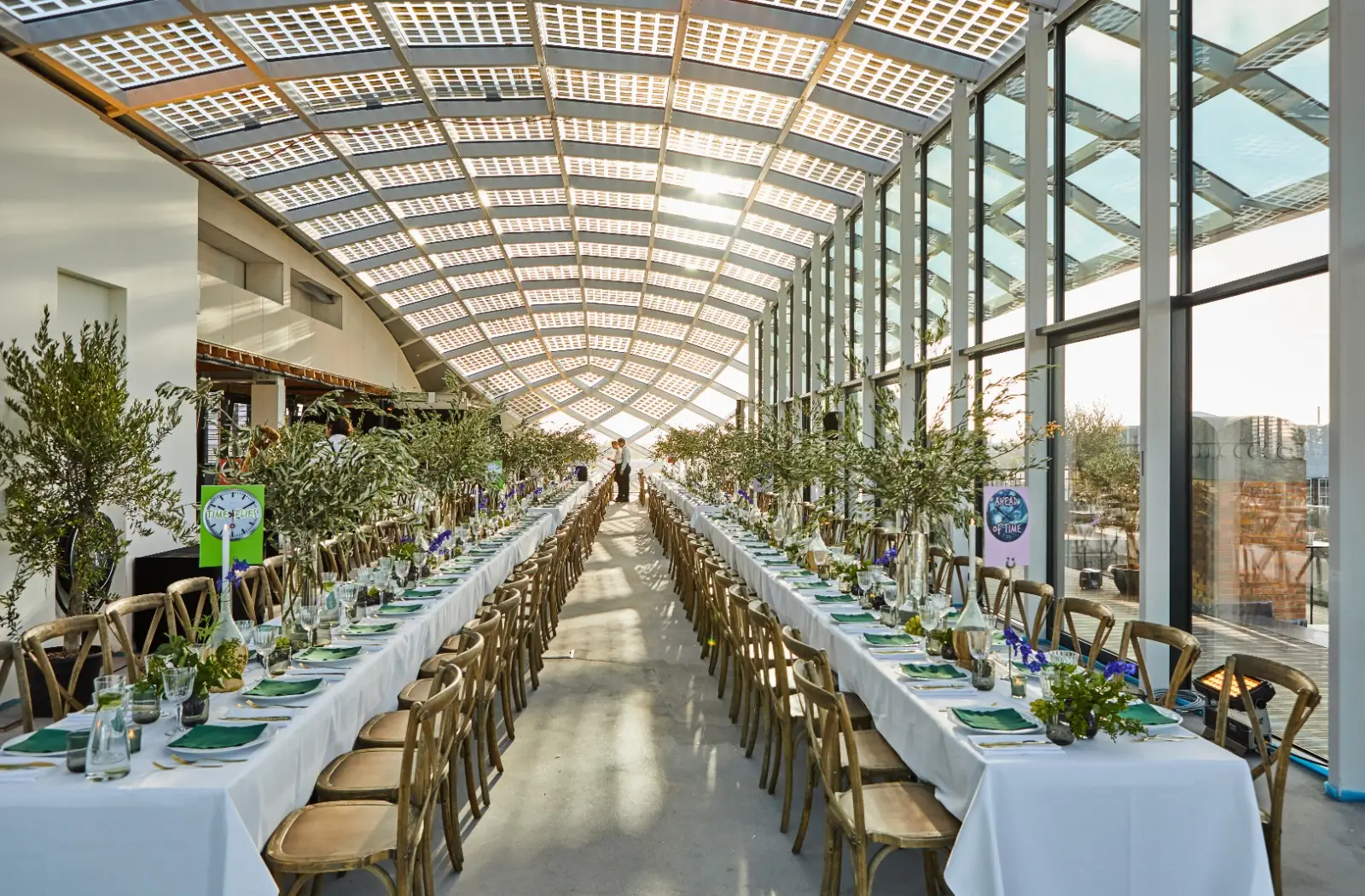 Long banquet setup in a glass atrium with two parallel rows of tables, greenery, and place settings ready for guests at sunset.