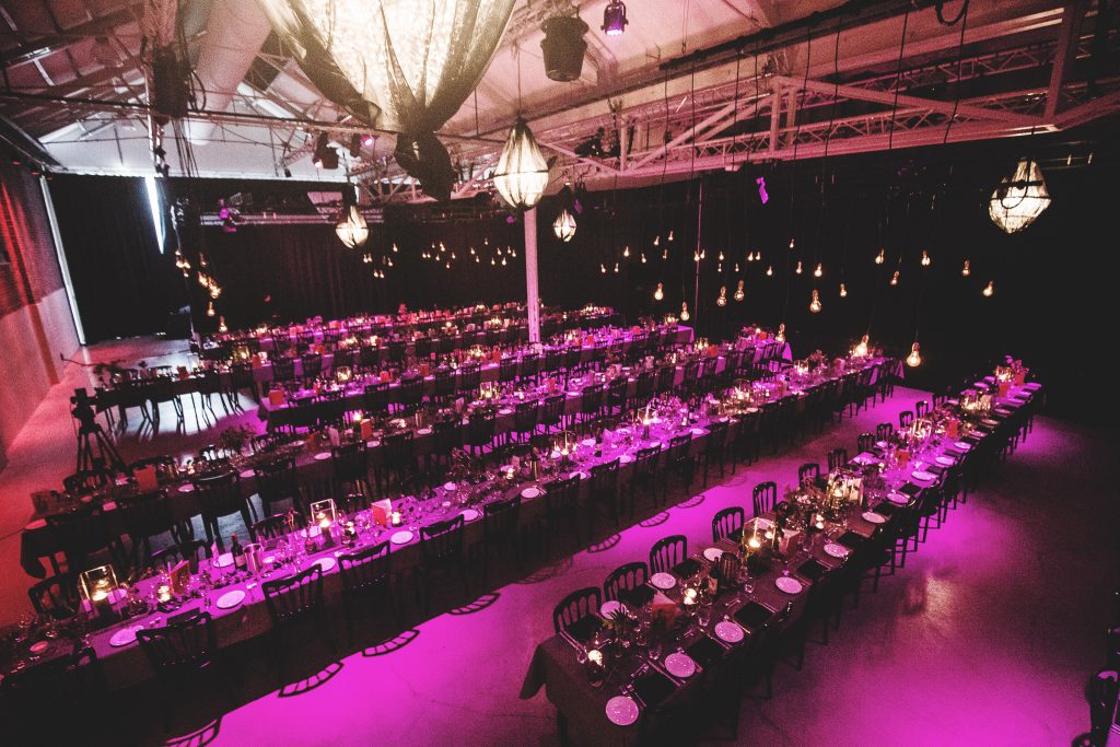 Long banquet tables set for a formal event in a dimly lit venue with pink-purple lighting and hanging pendant lights in the ceiling.