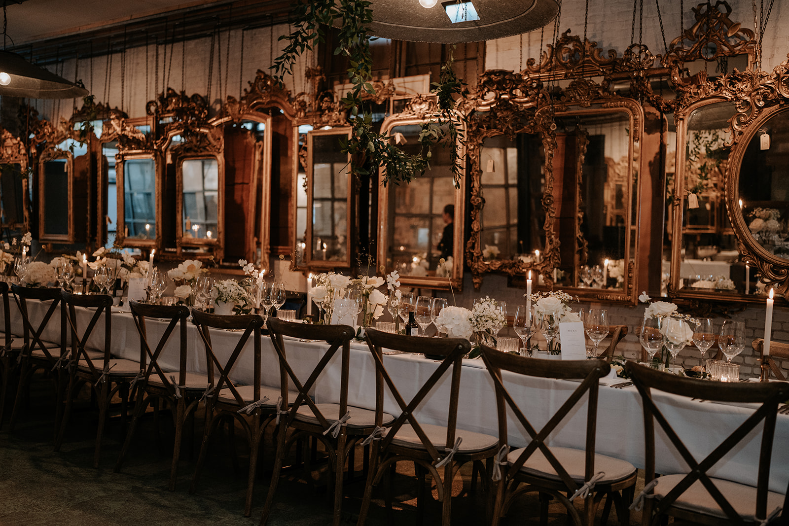 Long dining table set for a formal dinner with white flowers, candles, glassware, and vintage gilded mirrors along the wall.