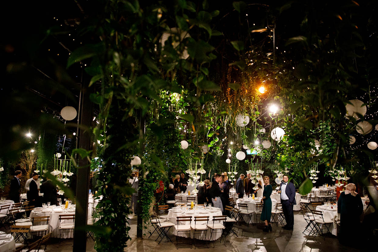 Guests mingle at a formal reception under hanging greenery and white lanterns in a greenhouse-style venue, evening light.