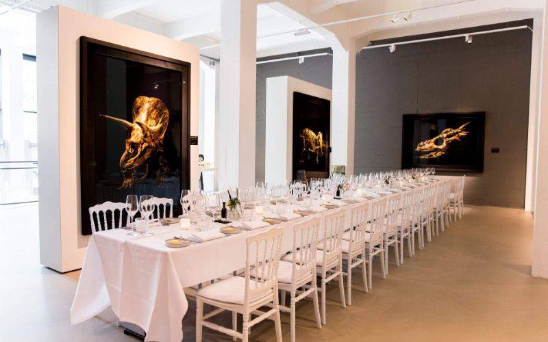 Long white-draped banquet table with white chairs in a gallery, set for a formal dinner among large artworks.