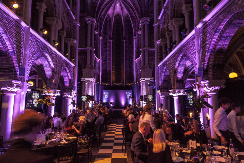 Guests seated at round tables for a formal dinner inside a vaulted brick hall lit in purple hues at a gala event