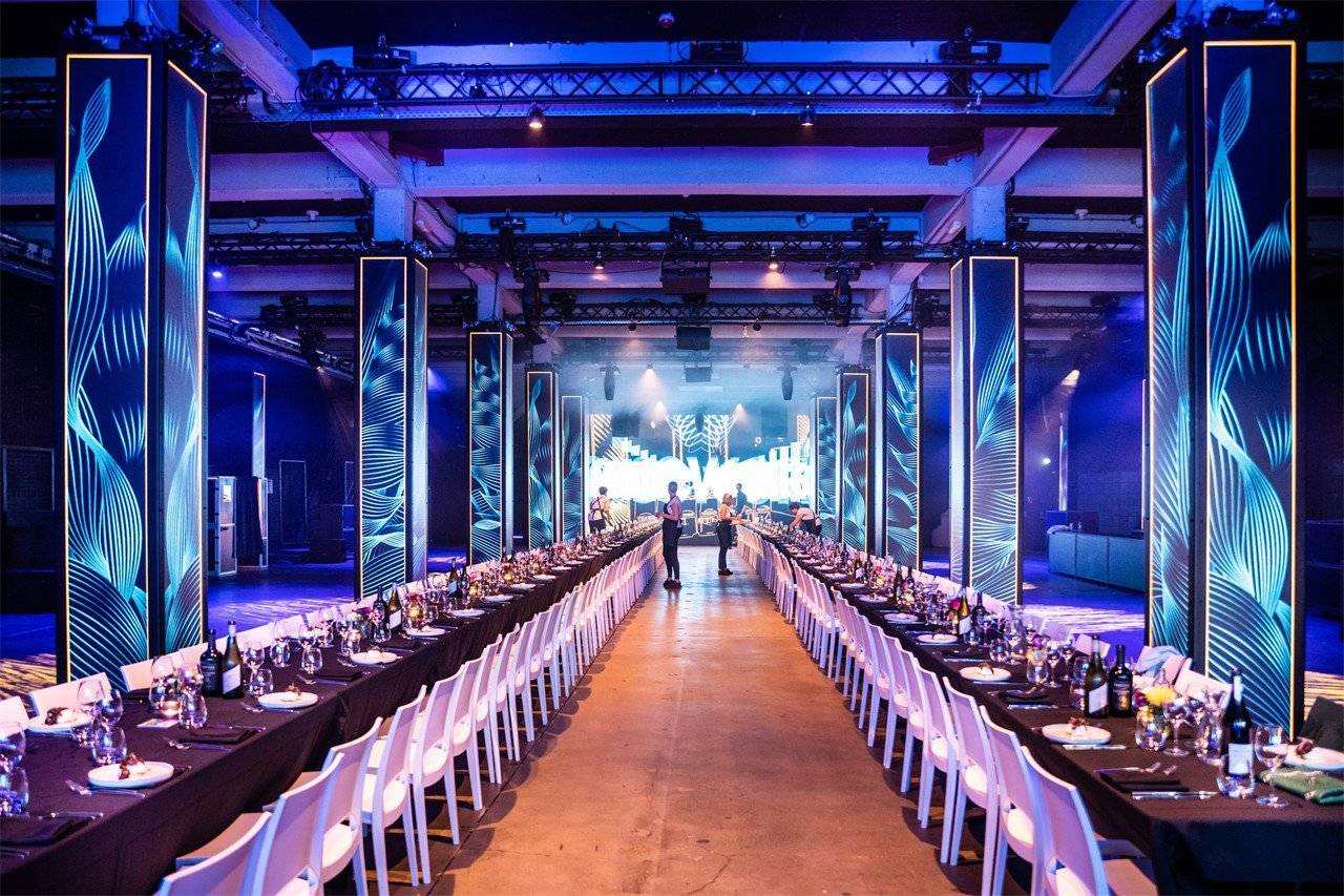 Banquet hall with long twin tables, white chairs, and blue-lit decorative pillars; people near the stage at the far end.