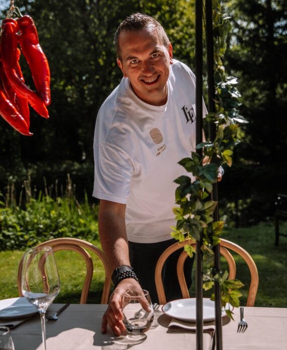 Smiling man in a white shirt at an outdoor dining table with wine glasses and plates; red peppers hang on the left.
