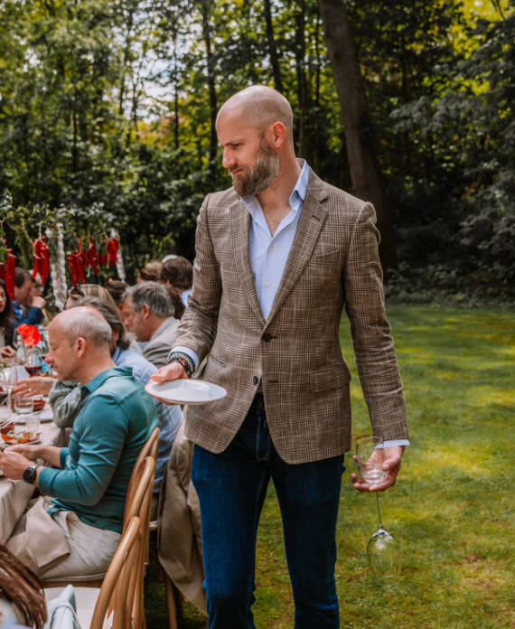 Man in a brown plaid blazer and blue shirt handing a plate to guests at an outdoor garden party.
