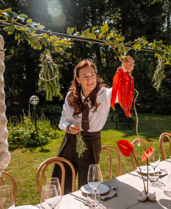 Woman outdoors hanging red peppers and herbs over a garden dining table set with glasses and plates.