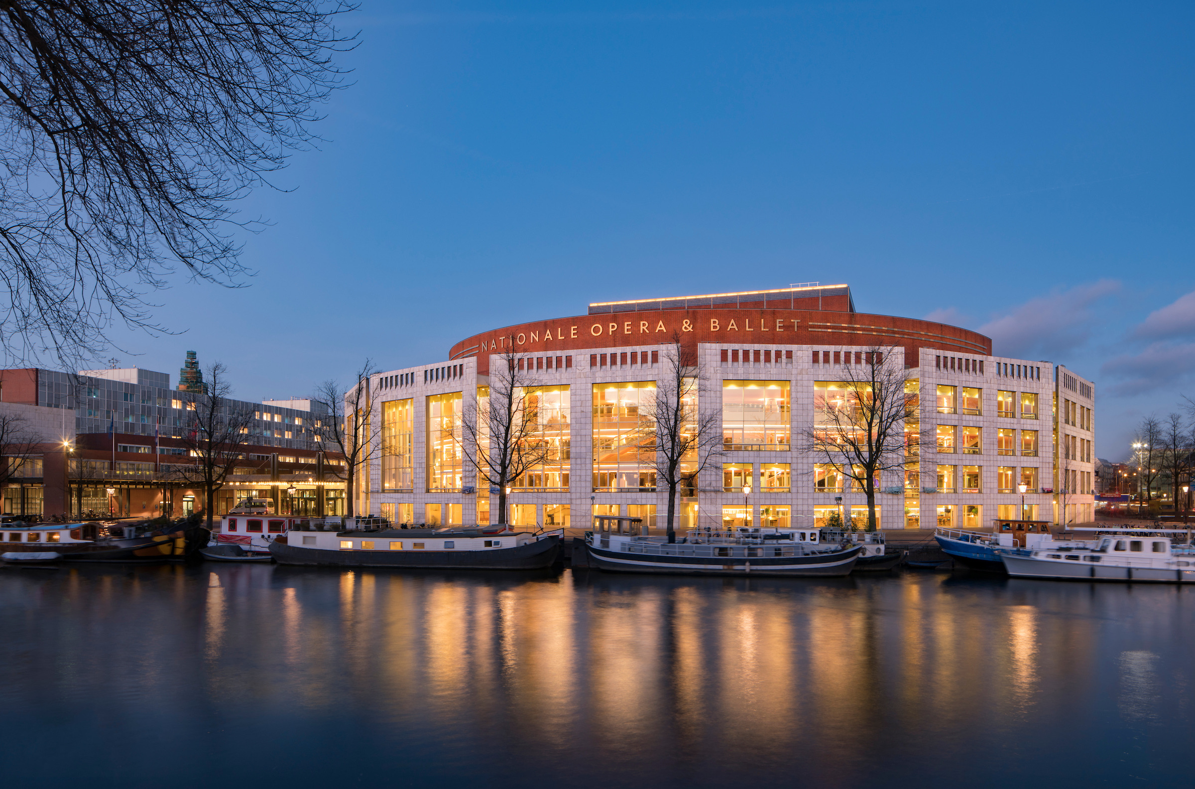 Night view of the National Opera & Ballet building along a calm river with warm-lit windows and boats docked nearby under a blue dusk sky = informative image informing about the venue and waterfront setting, 2-3 sentences? Wait need concise.