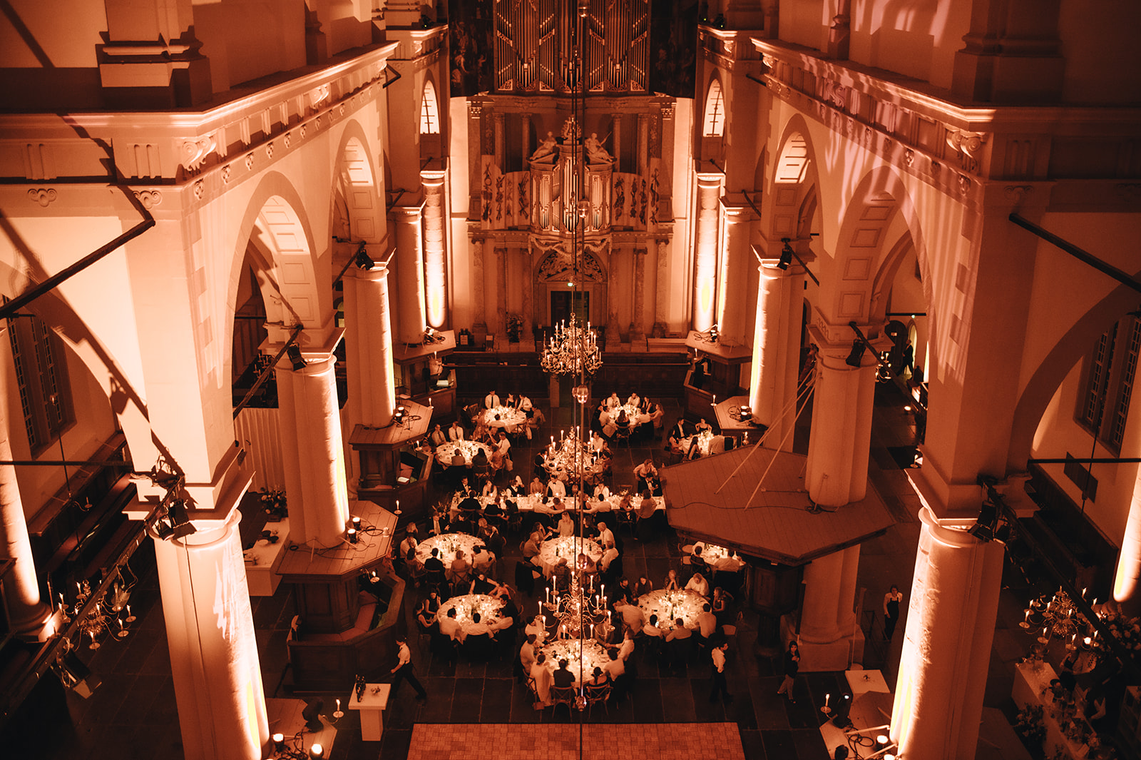 Ornate hall set for dinner: high arches, columns, chandeliers, and guests at round tables in warm orange lighting.