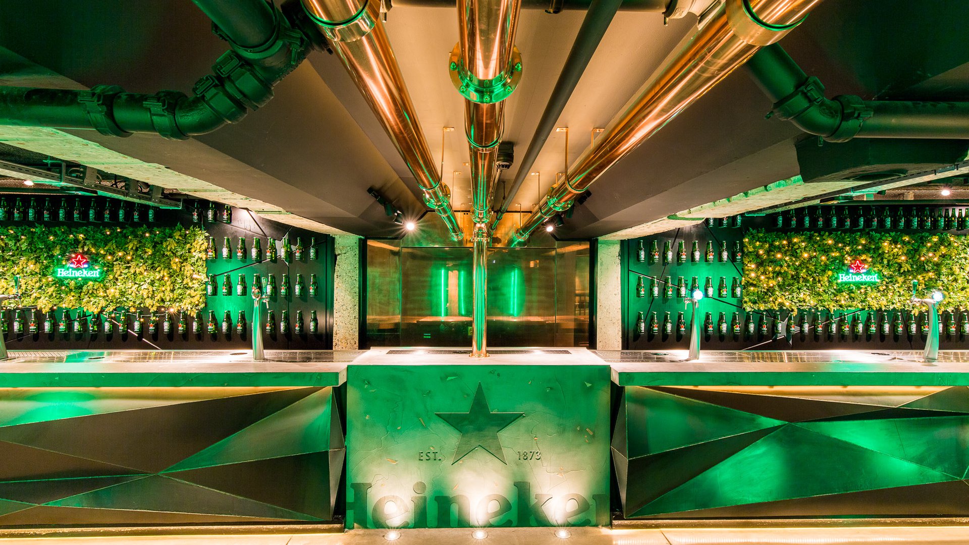 Indoor bar display with rows of Heineken bottles on ivy-covered walls and green neon Heineken signs.