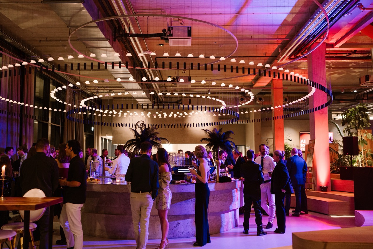 Guests mingle around a curved bar at a stylish indoor party with circular hanging lights and pink-purple mood lighting.