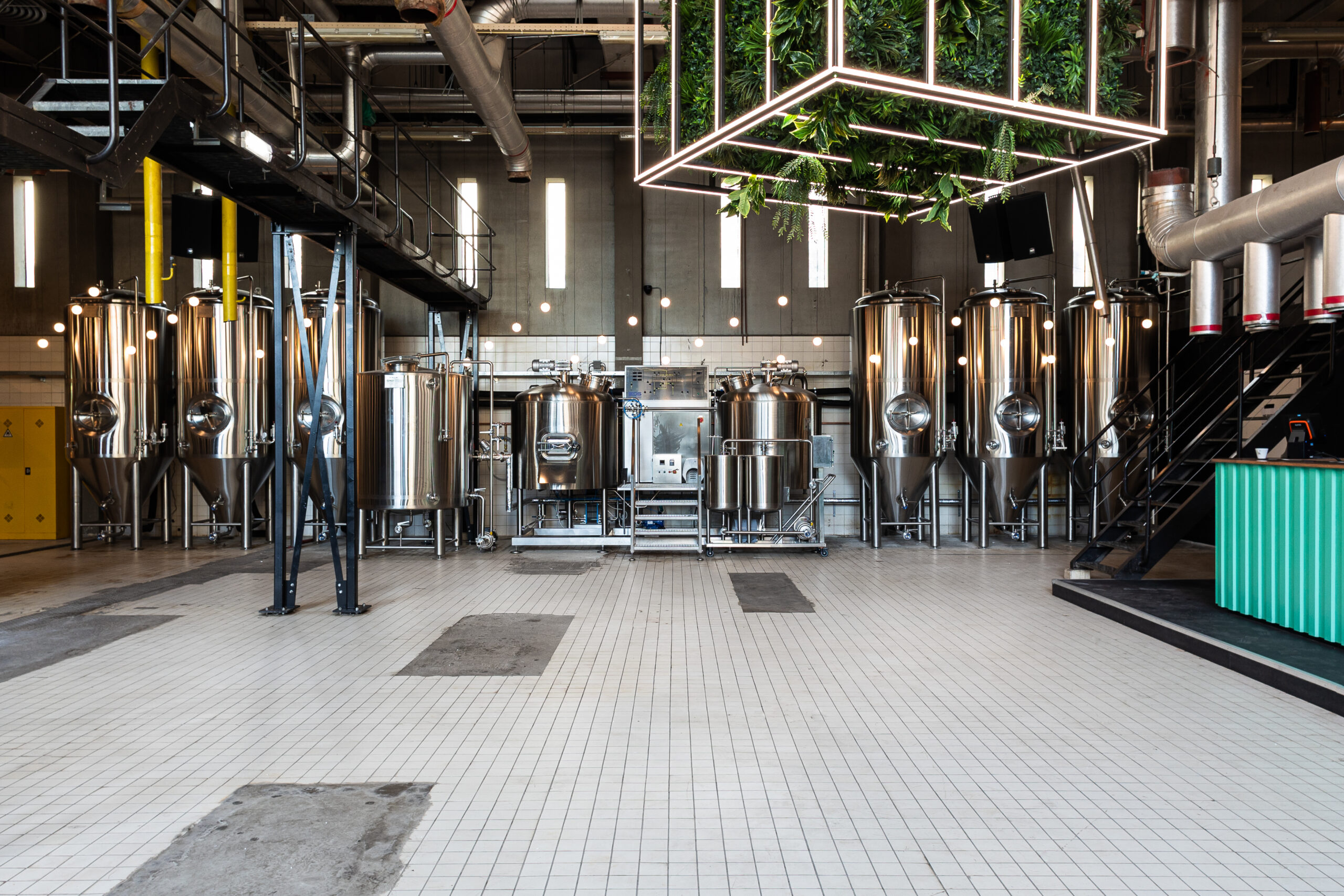 Industrial brewery interior with large shiny stainless steel fermenters and metal piping along walls and ceiling, tiled floor beneath.