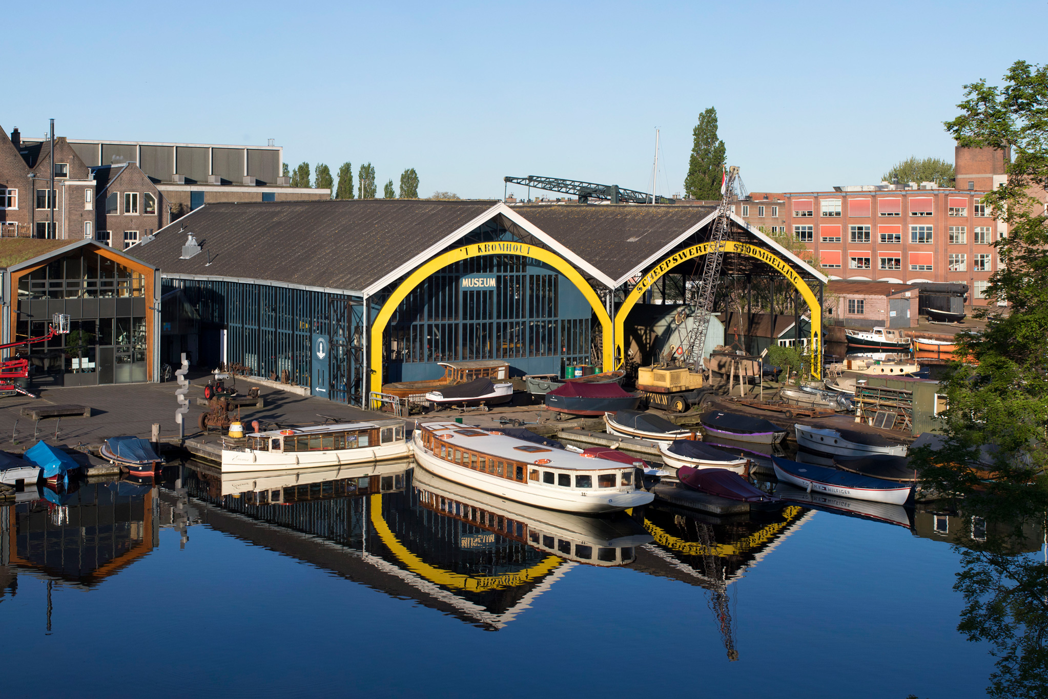 Marina scene with moored boats and a glass-fronted boathouse framed by bright yellow arches against a blue sky and brick buildings behind.