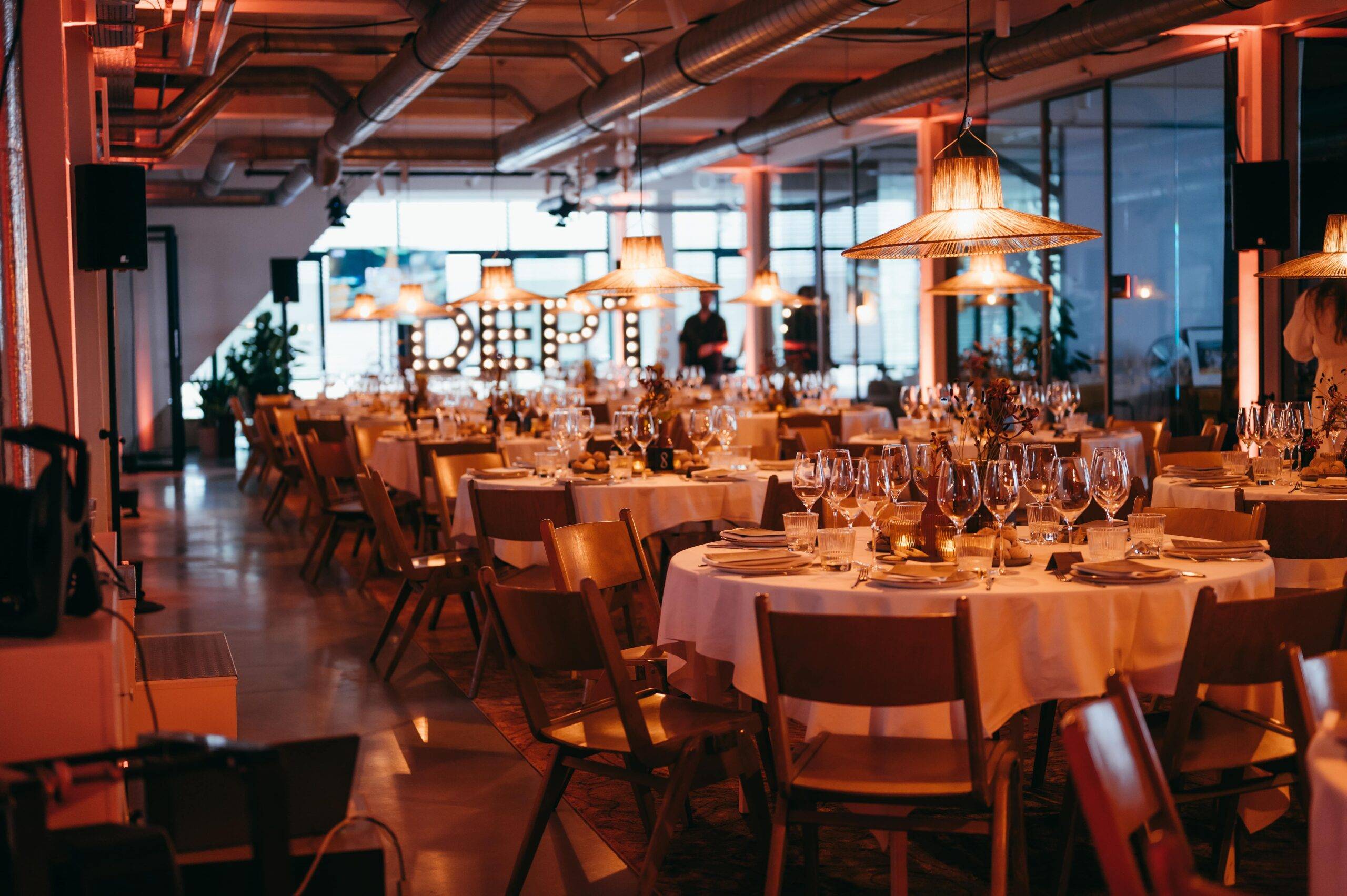 Banquet hall set for a dinner with round tables, white tablecloths, wine glasses, and warm pendant lighting.