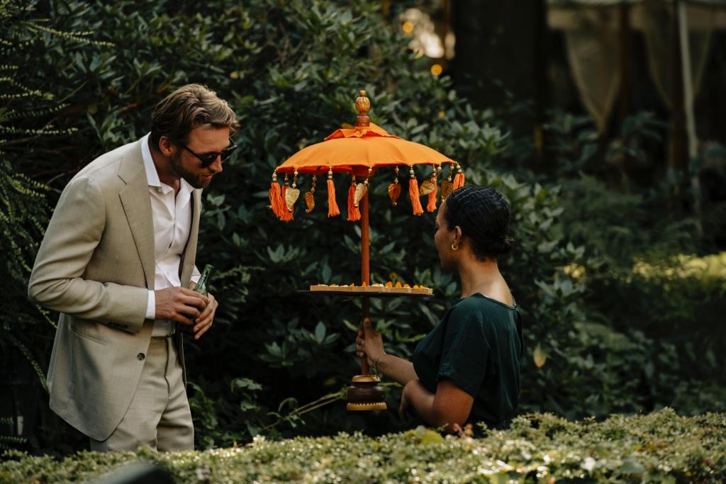 Man in a light beige suit and sunglasses chats with a woman holding a tray under an orange tassel-bordered parasol outdoors, with treats on the tray.
