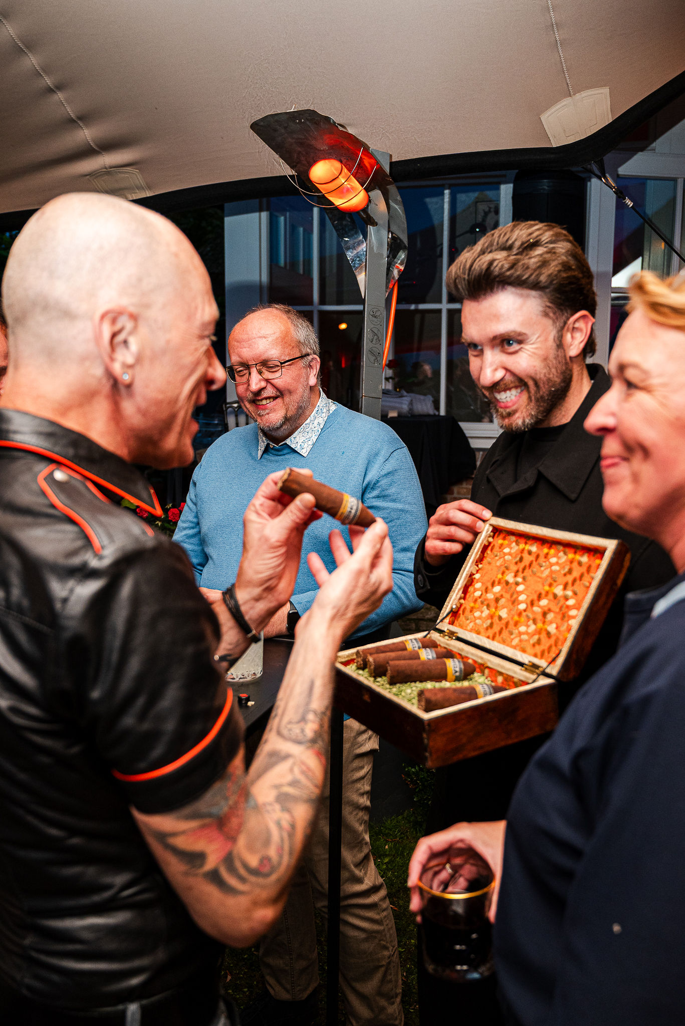 Group of adults at a casual outdoor party, several cigars in hand and an open box of cigars on the table