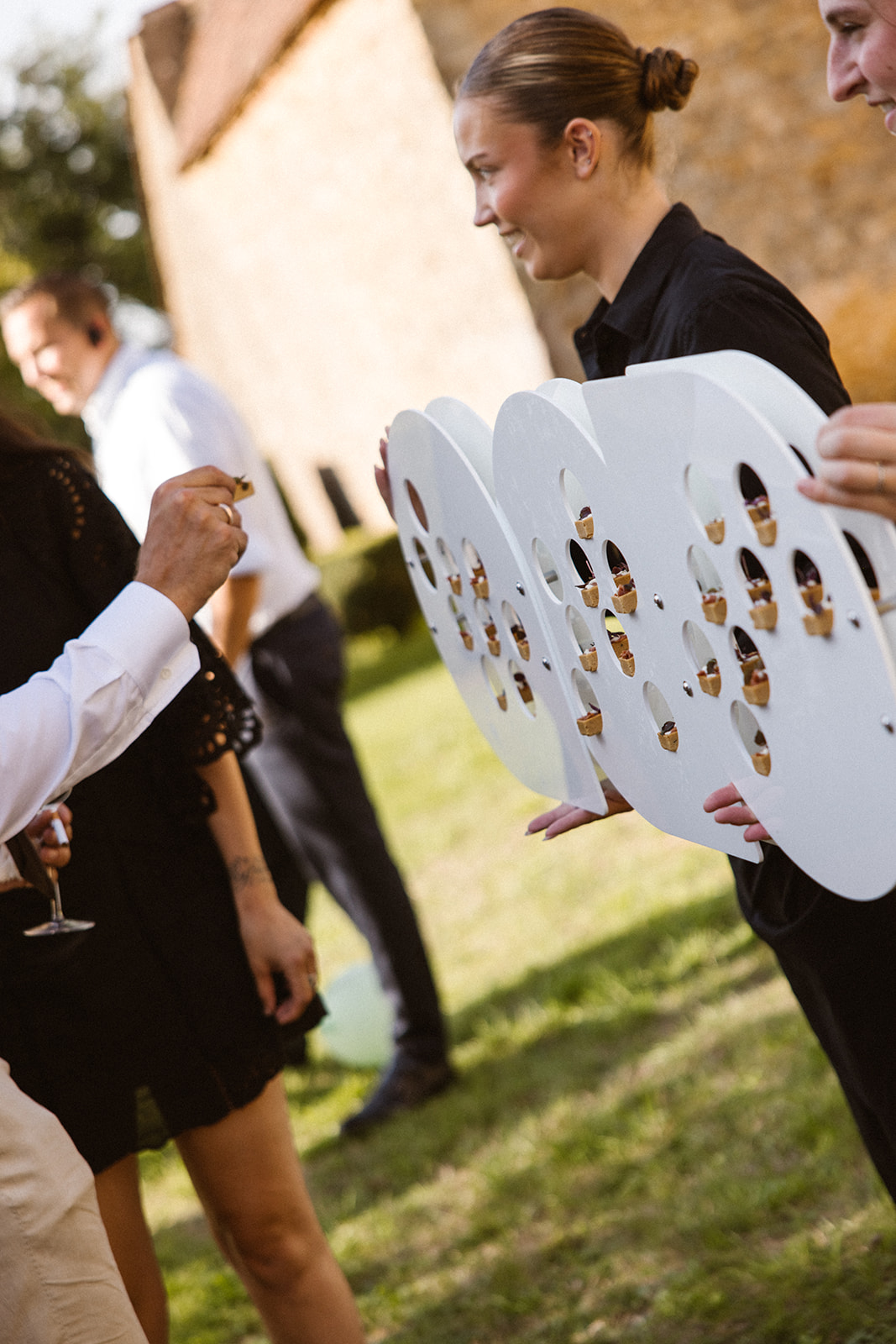 Staff in black hold large white circular trays filled with bite-sized desserts, serving a guest at an outdoor event.
