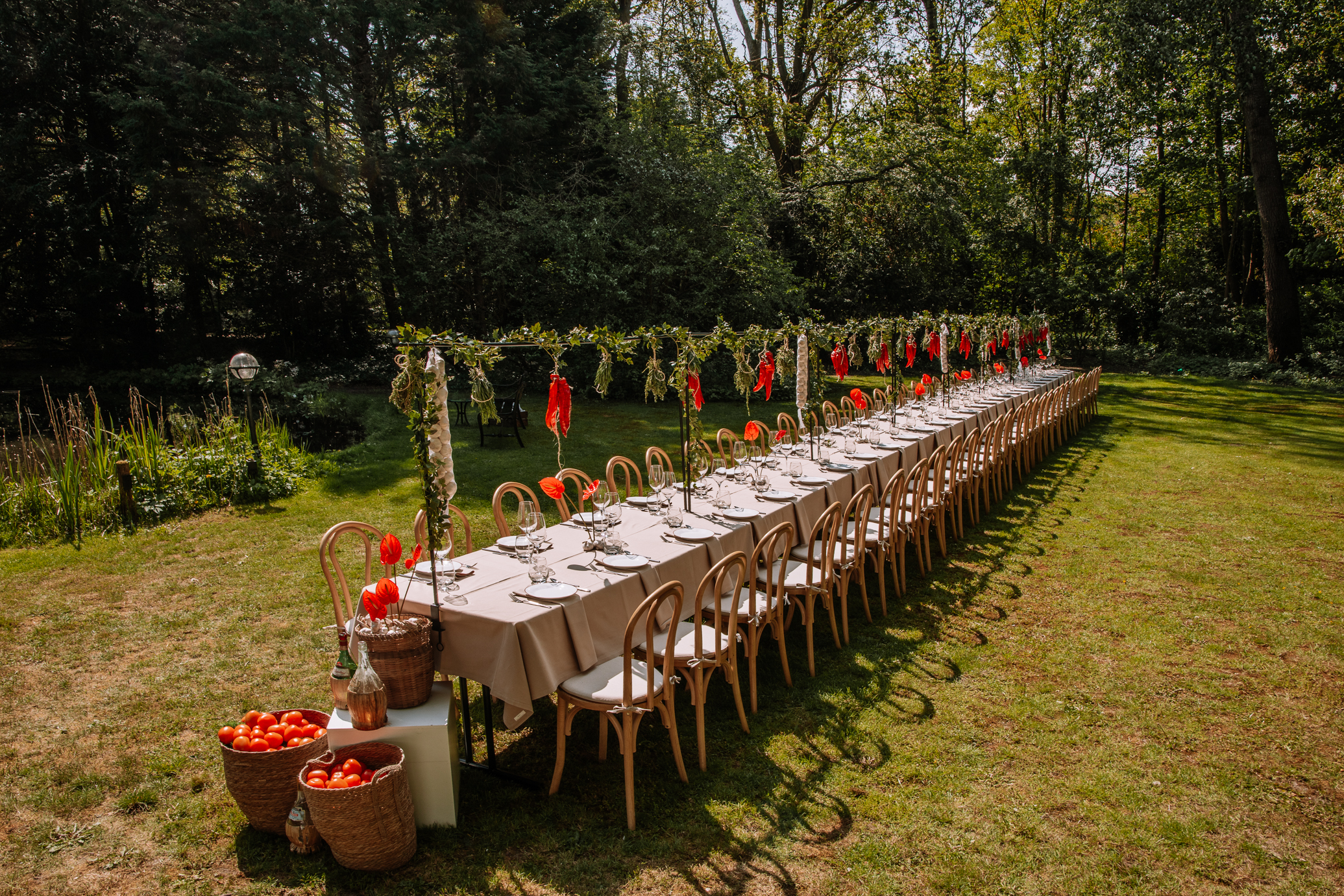 Long outdoor garden dining setup with a long white-clothed table, wooden chairs, greenery garlands, and red decorations for an event.