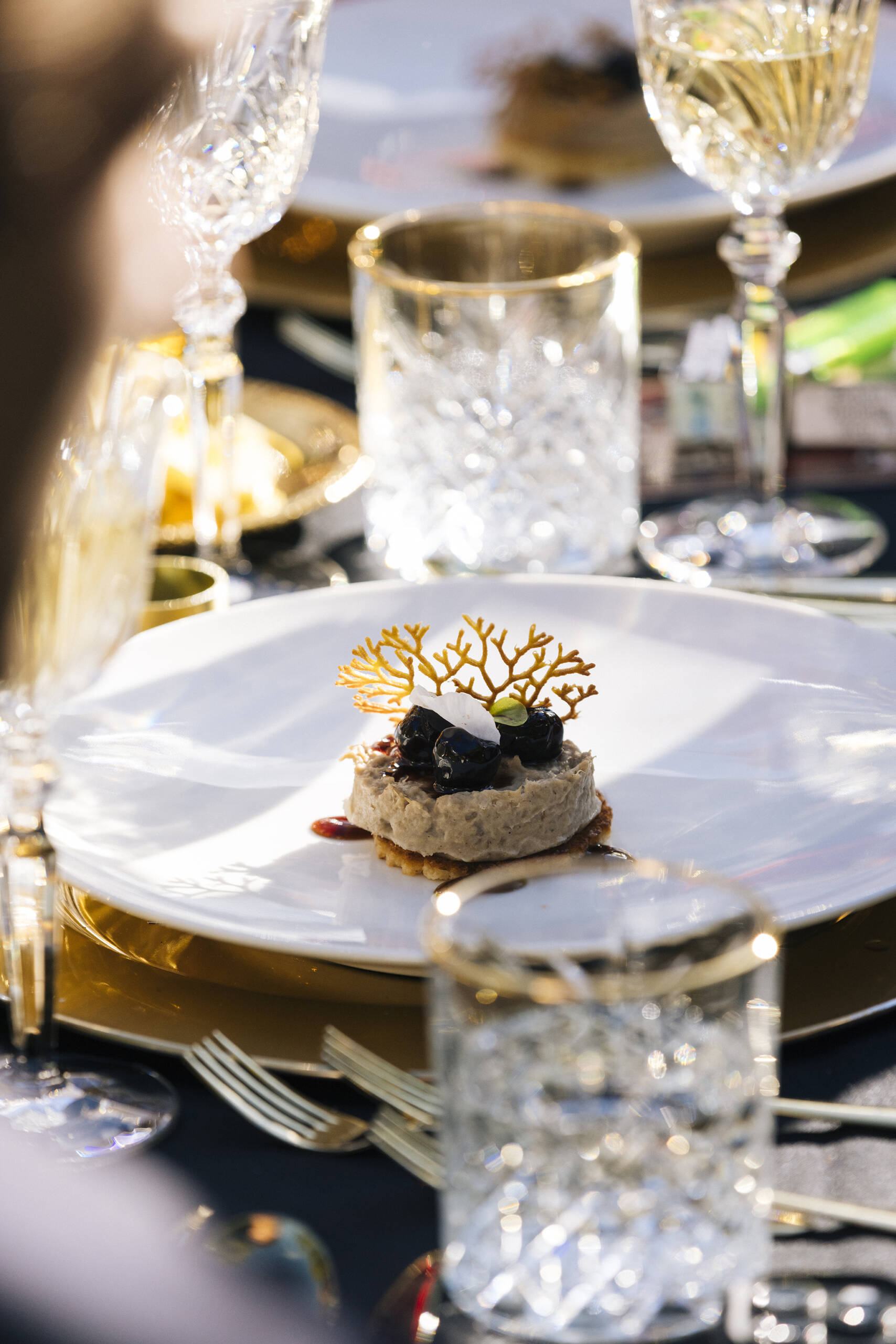 Dessert on a white plate with blueberries, cream, and a coral sugar garnish, set on a gold charger with crystal glasses nearby.
