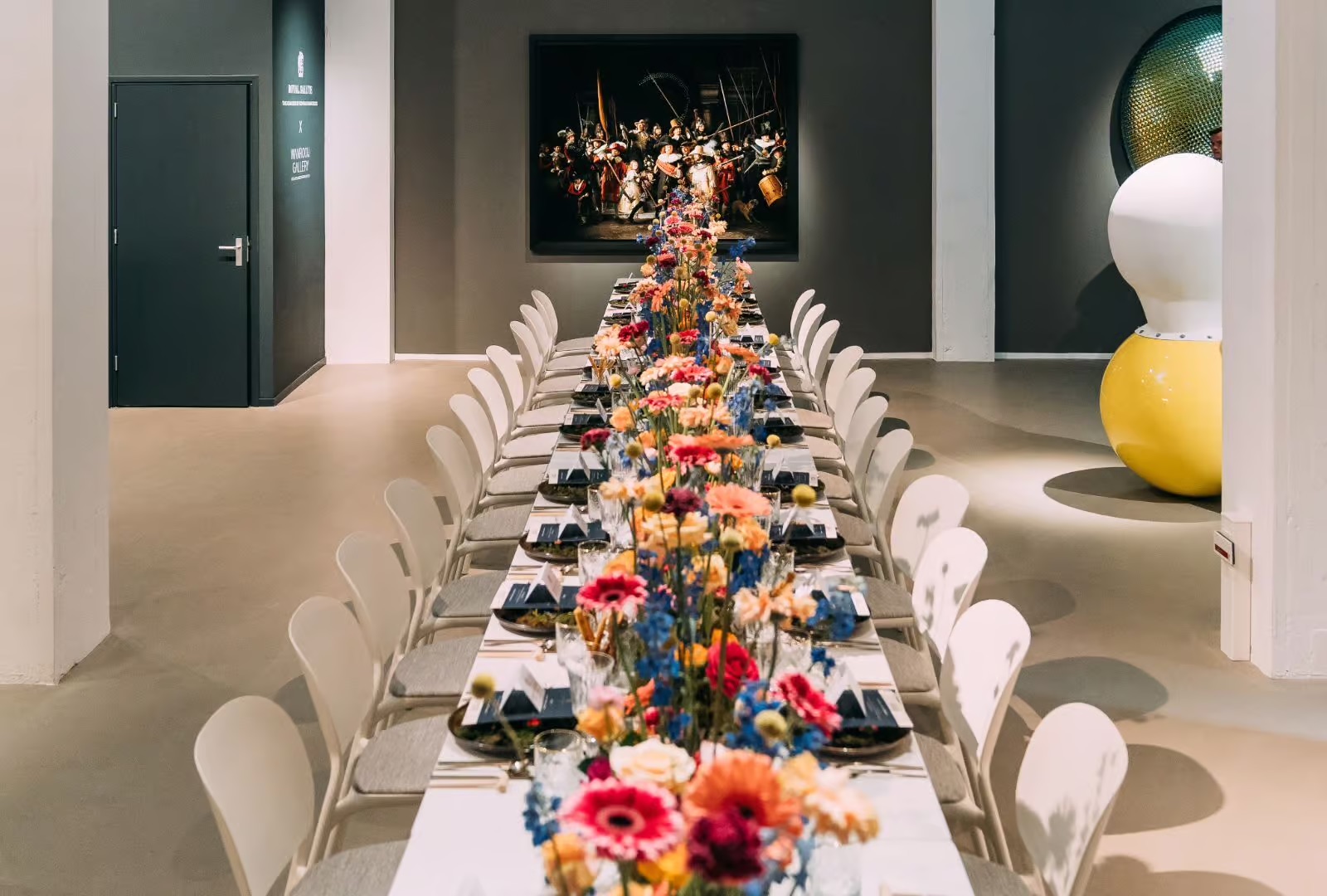 Long banquet table set for a formal dinner with colorful flower centerpieces in a gallery setting, with artwork on the back wall and a sculpture nearby.