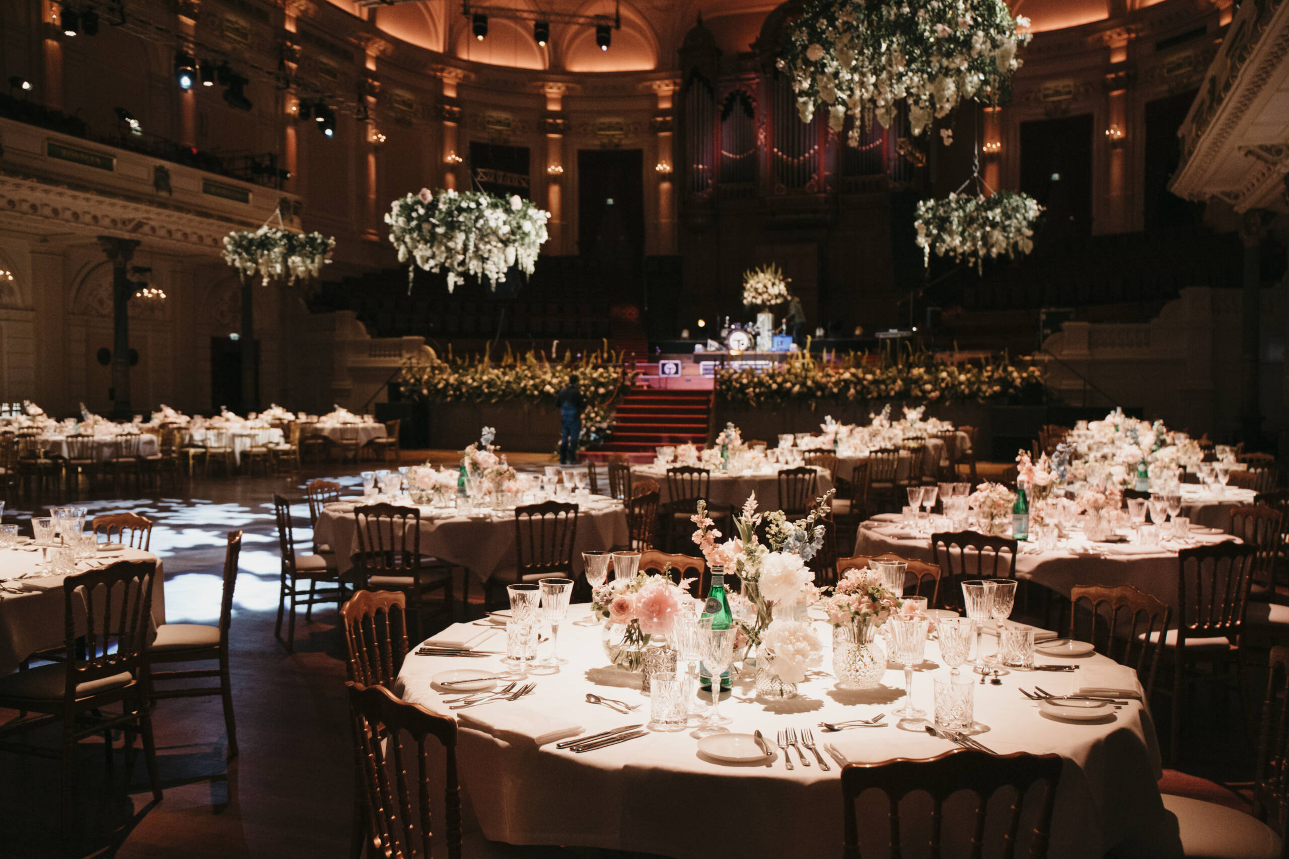 Elegant ballroom banquet setup with round tables, white linens, glassware, and tall floral centerpieces under warm chandeliers, stage in back.