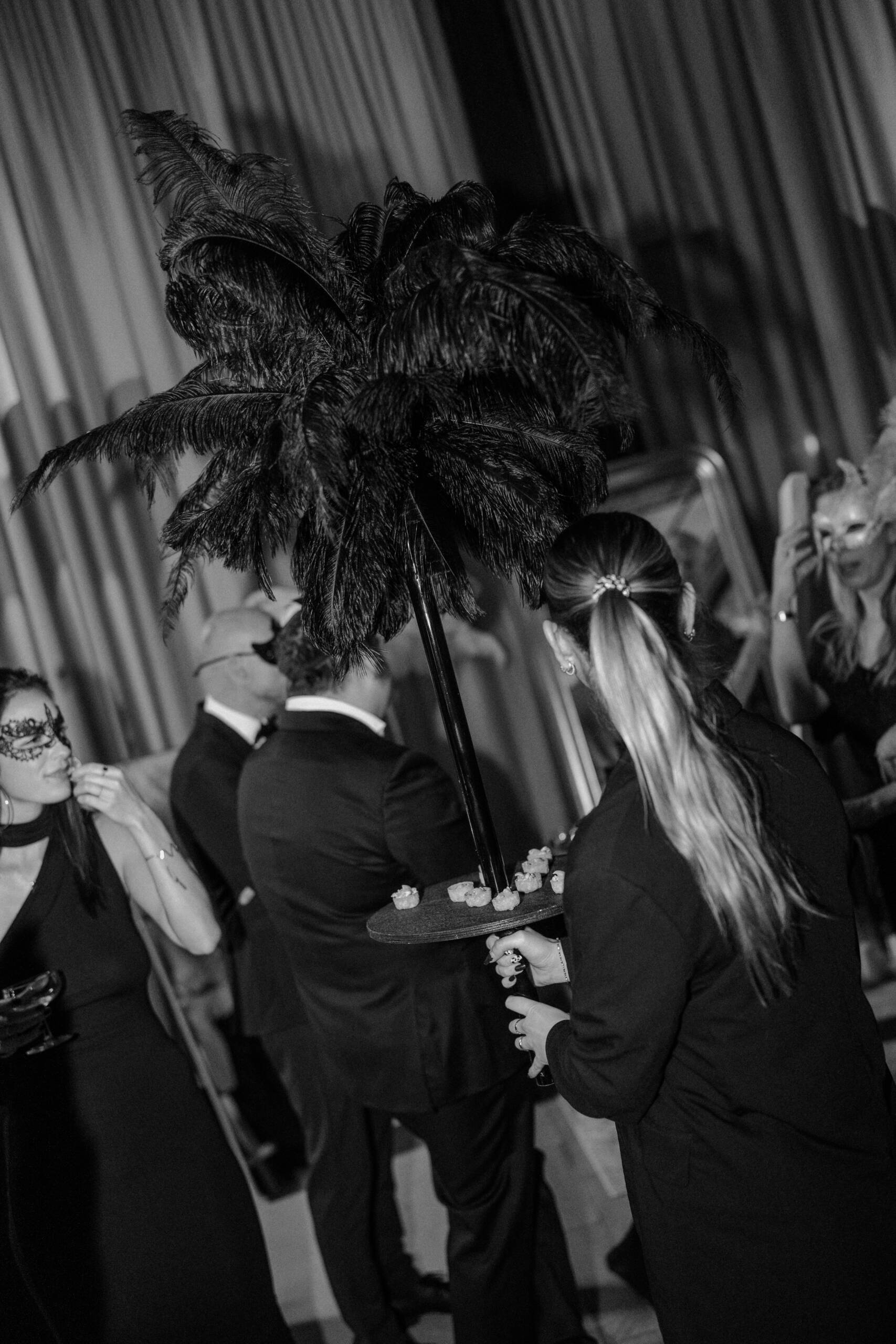 Formal black-and-white party scene with guests in evening wear; a tall feather centerpiece sits on a tray of hors d'oeuvres held by a guest in the foreground.