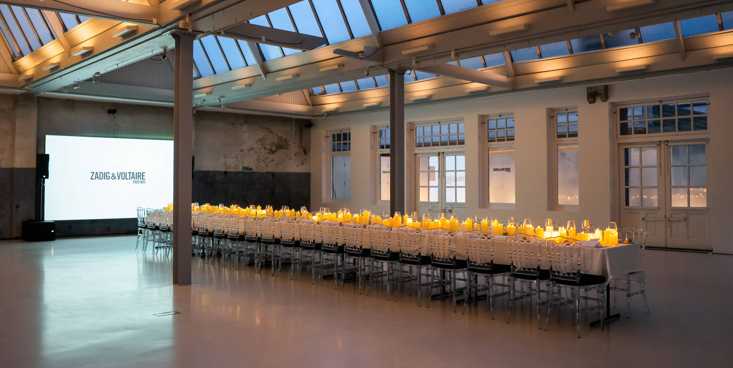 Long candle-lit banquet table in a skylit industrial venue, with a projection screen reading Zadig & Voltaire.