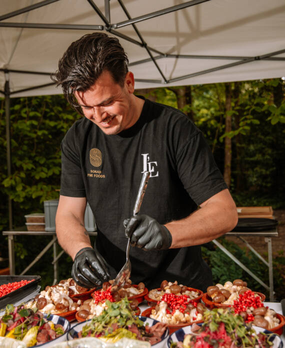 Man in black t-shirt and gloves serving colorful hors d'oeuvres under a canopy at an outdoor setup.