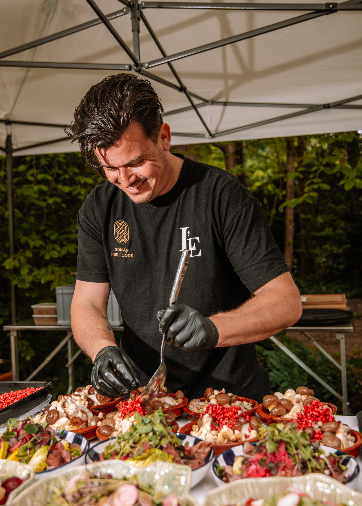 Man wearing black T-shirt and gloves plating dessert bowls under a white canopy, using tongs and a spoon.