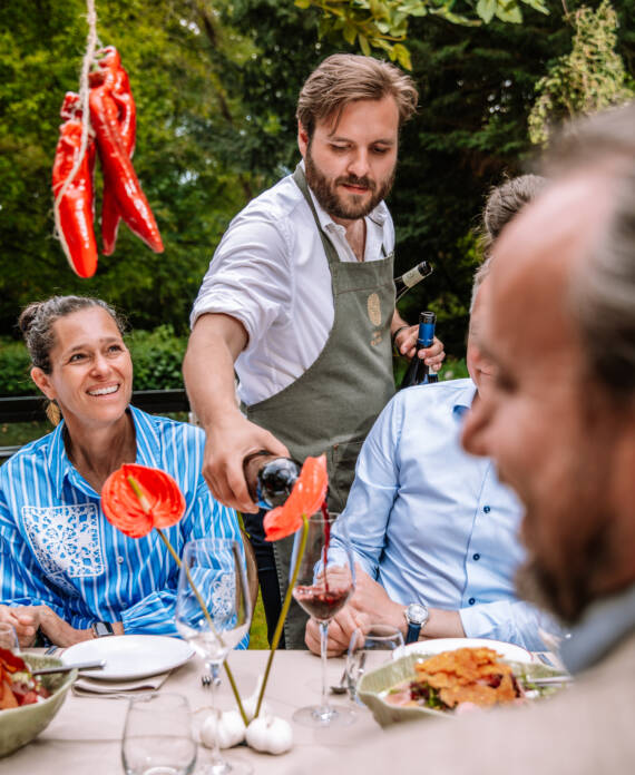 Bearded man in a gray apron pours wine for a group at an outdoor meal, with red peppers hanging above. (Outdoor garden party)