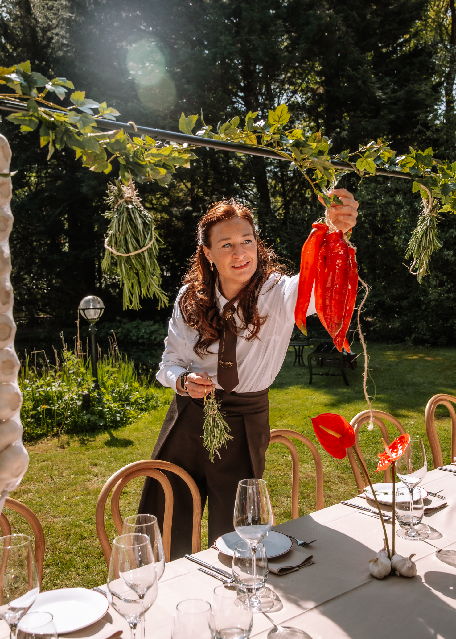 Woman in a white blouse and brown apron hangs bright red peppers and herbs from a string over an outdoor dinner table set with glassware and plates.