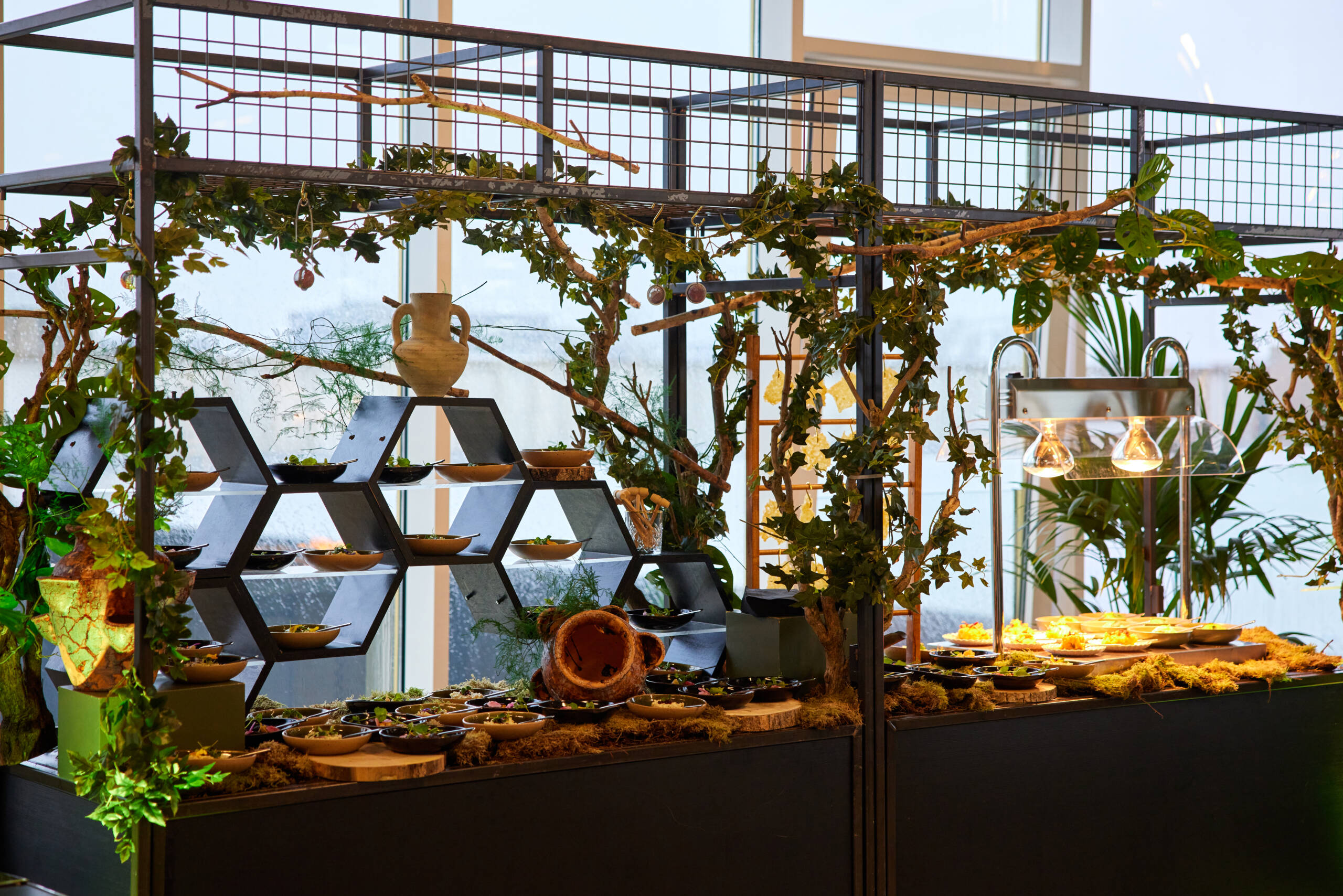 Plant-filled buffet area in a restaurant with leafy vines, ceramic bowls, and geometric shelves.