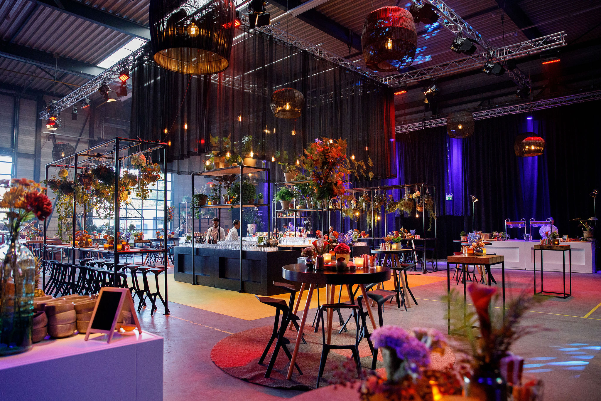 Indoor event hall with a long buffet bar, flower arrangements on metal stands, and dramatic hanging lights suspended from a truss system under black drapes.