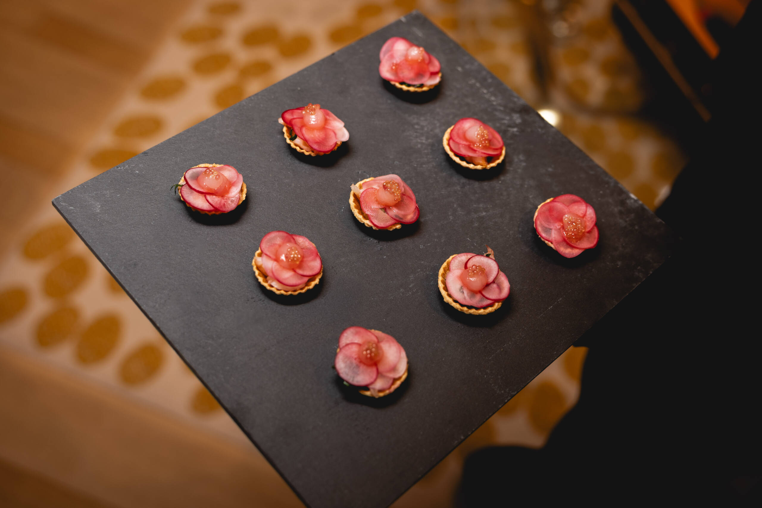Small round pastry bites topped with pink flower-like petals on a dark slate serving board.