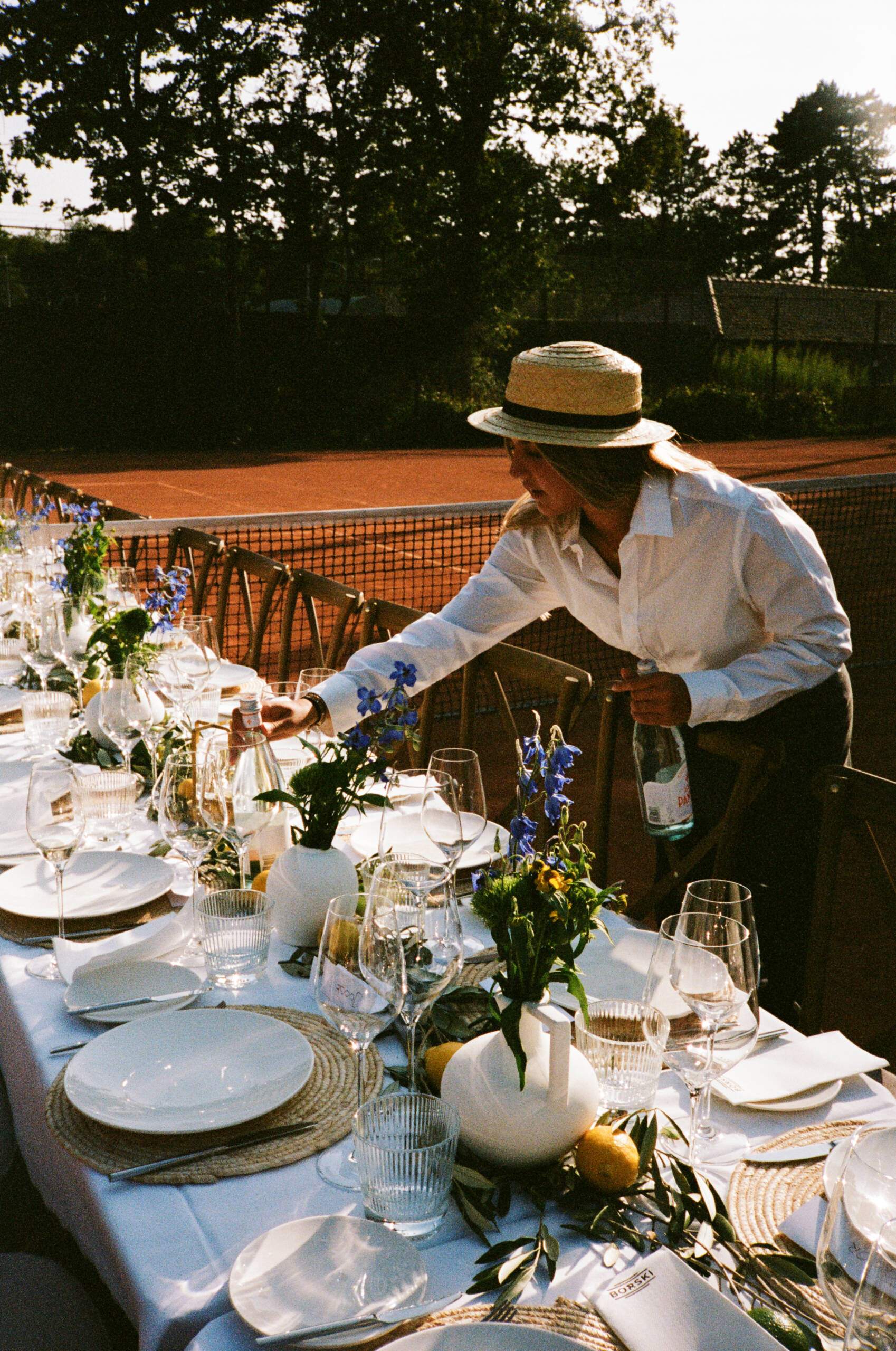 Woman in a white shirt and straw hat leaning to arrange a long outdoor dinner table with wine glasses, plates, and blue floral centerpieces on a sunny terrace behind a tennis court.