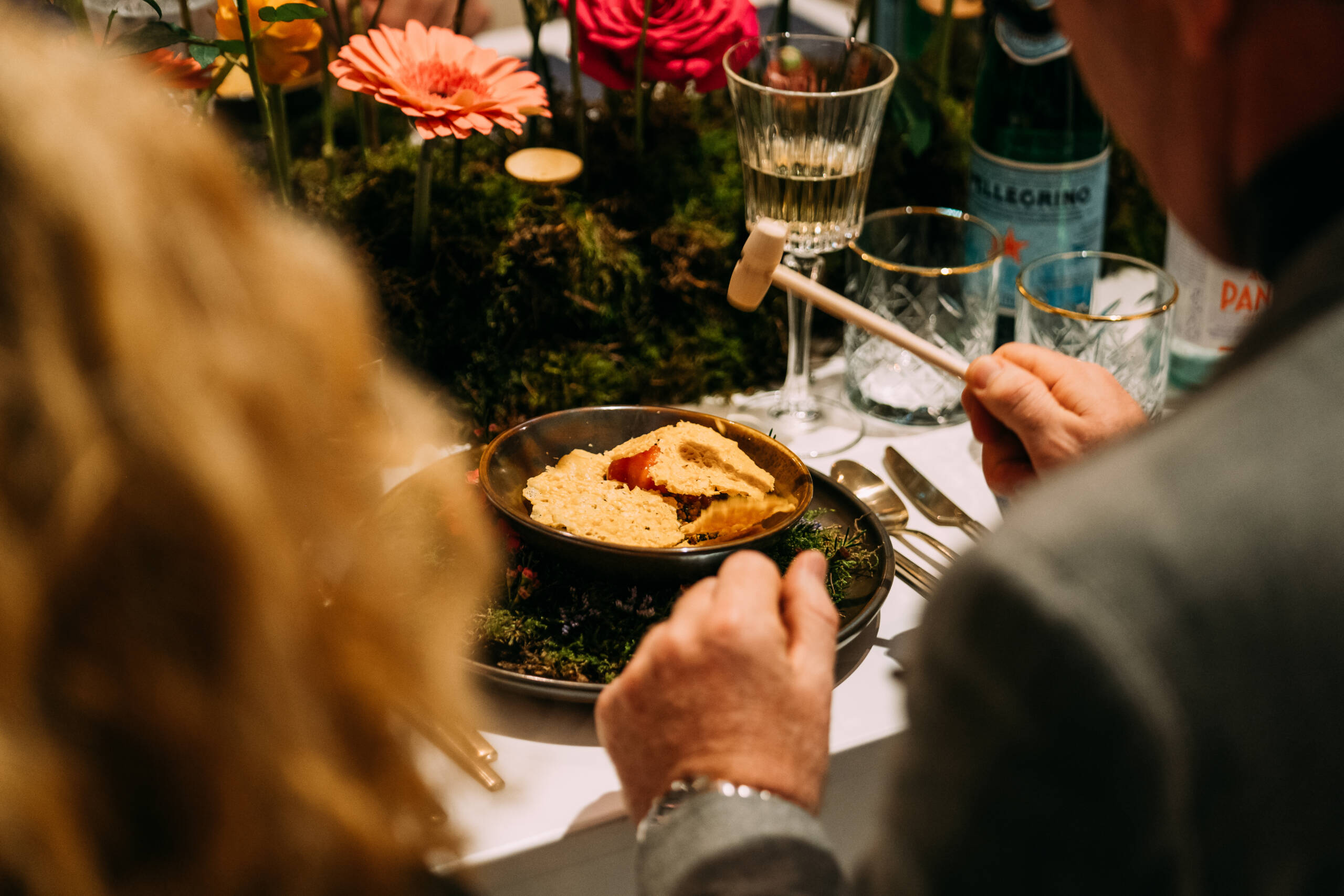 People seated around a table share a pie, with a hand reaching in to serve a slice as flowers, glassware, and a bottle appear in the background.