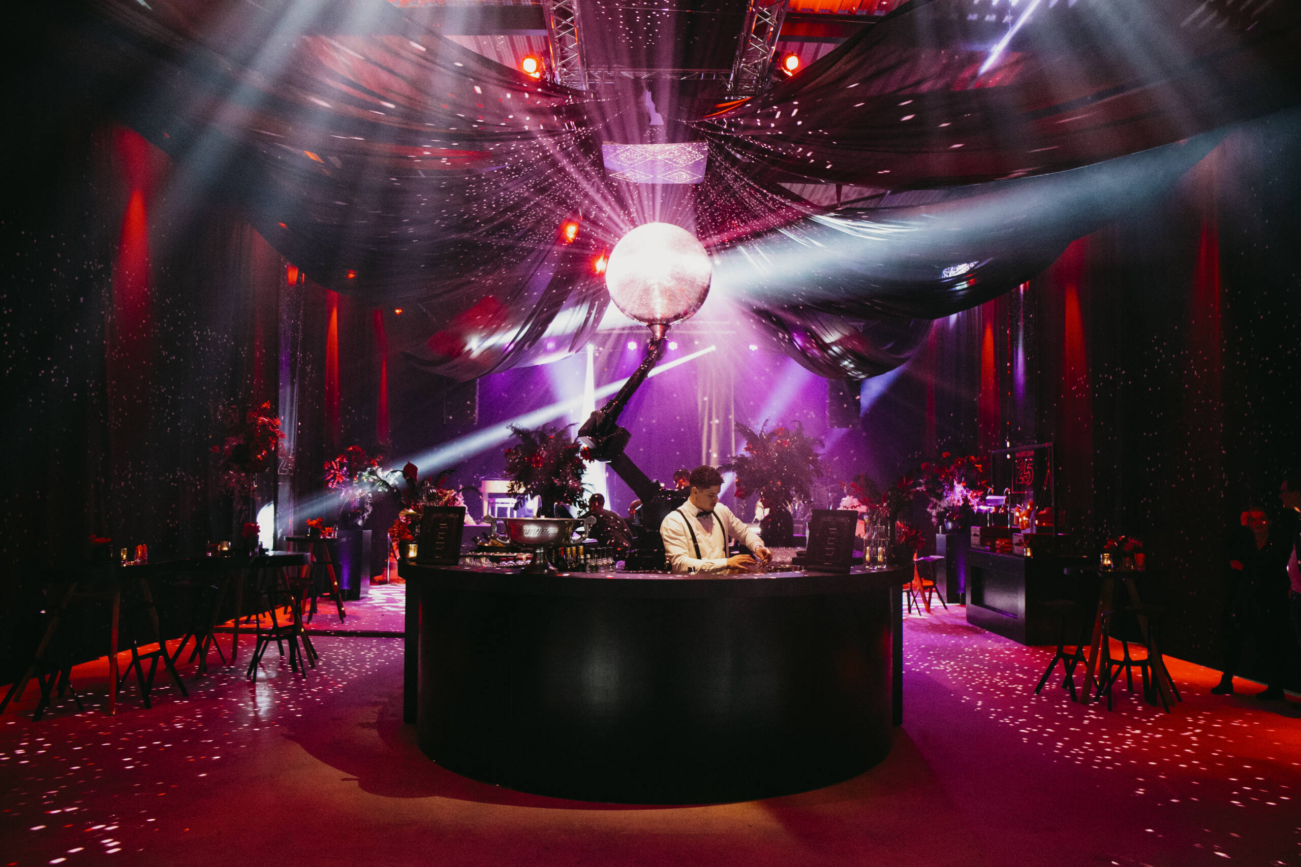 DJ booth in a dark club with a glittering disco ball, red and purple lights, and a bartender mixing drinks at the curved bar