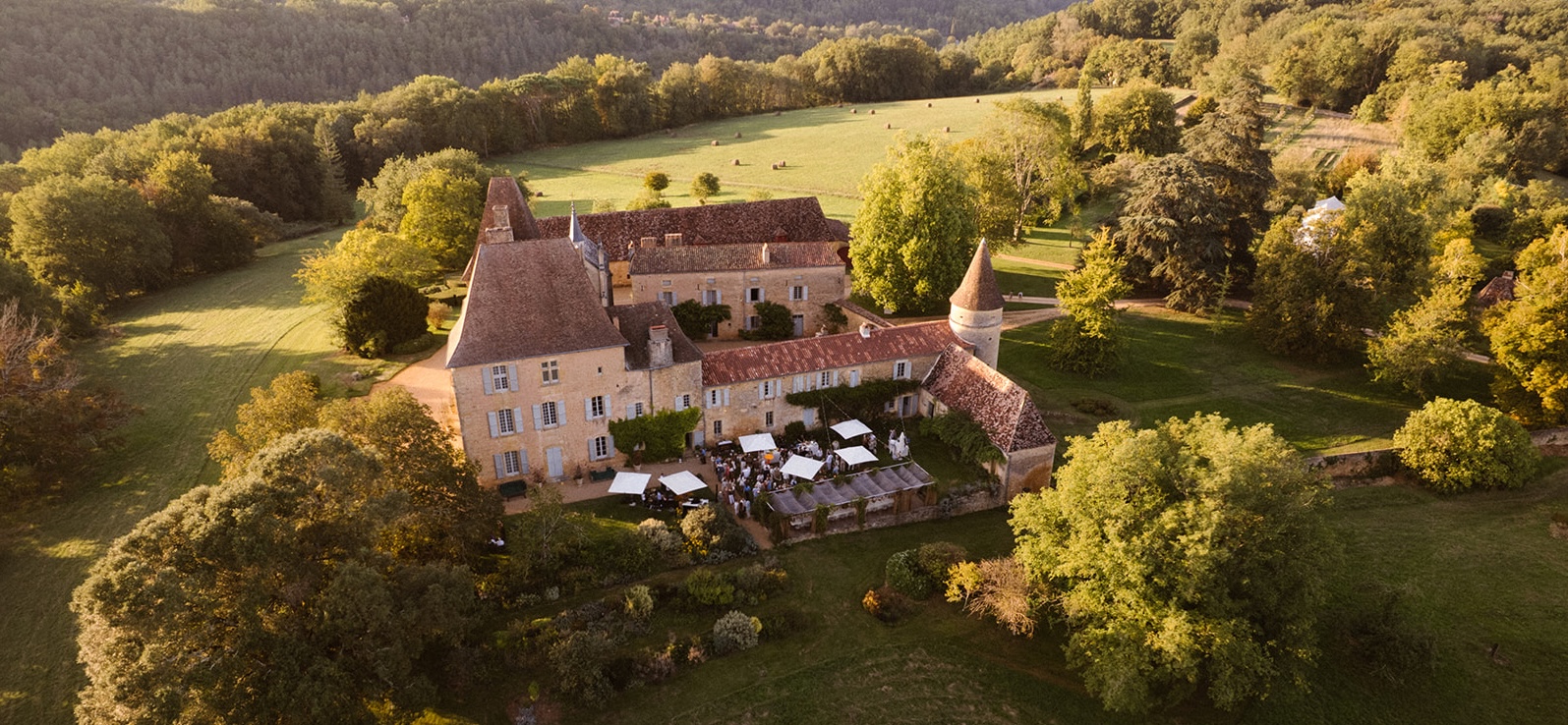 Aerial view of a historic stone castle with red-tiled roofs hosting an outdoor gathering under white canopy tents in a green landscape.