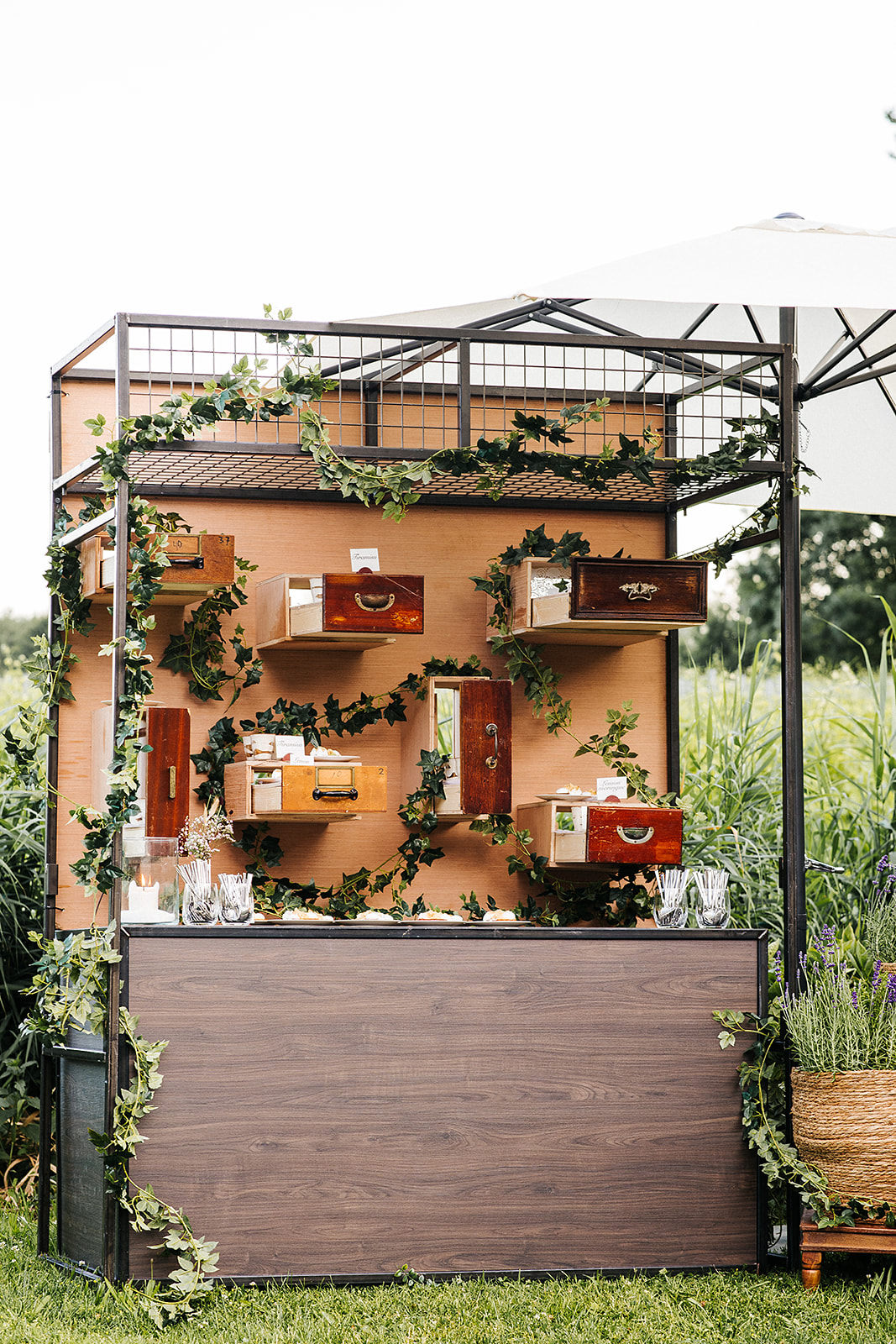 Outdoor display booth with stacked vintage wooden drawers arranged on a black metal frame, ivy garlands wrapping around the structure, and a cream umbrella overhead.