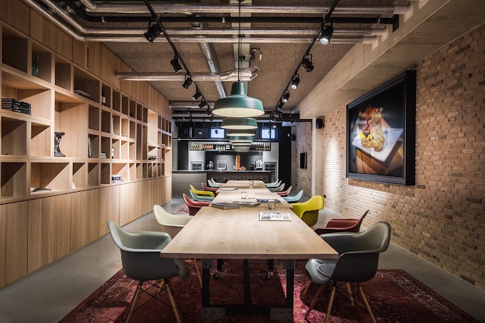 Long wooden conference table with colorful chairs in a modern office break room; brick walls, open shelves on the left, a kitchen at the far end, and a framed food image on the right.