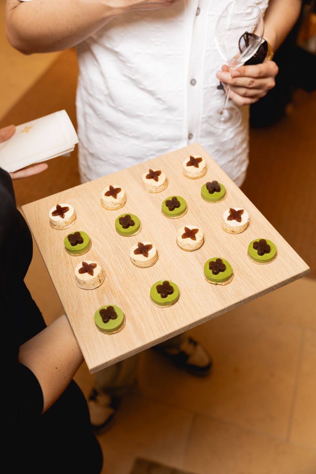 Tray of bite-sized desserts on a wooden board: green discs and beige rounds topped with dark chocolate shapes, being offered at a tasting.