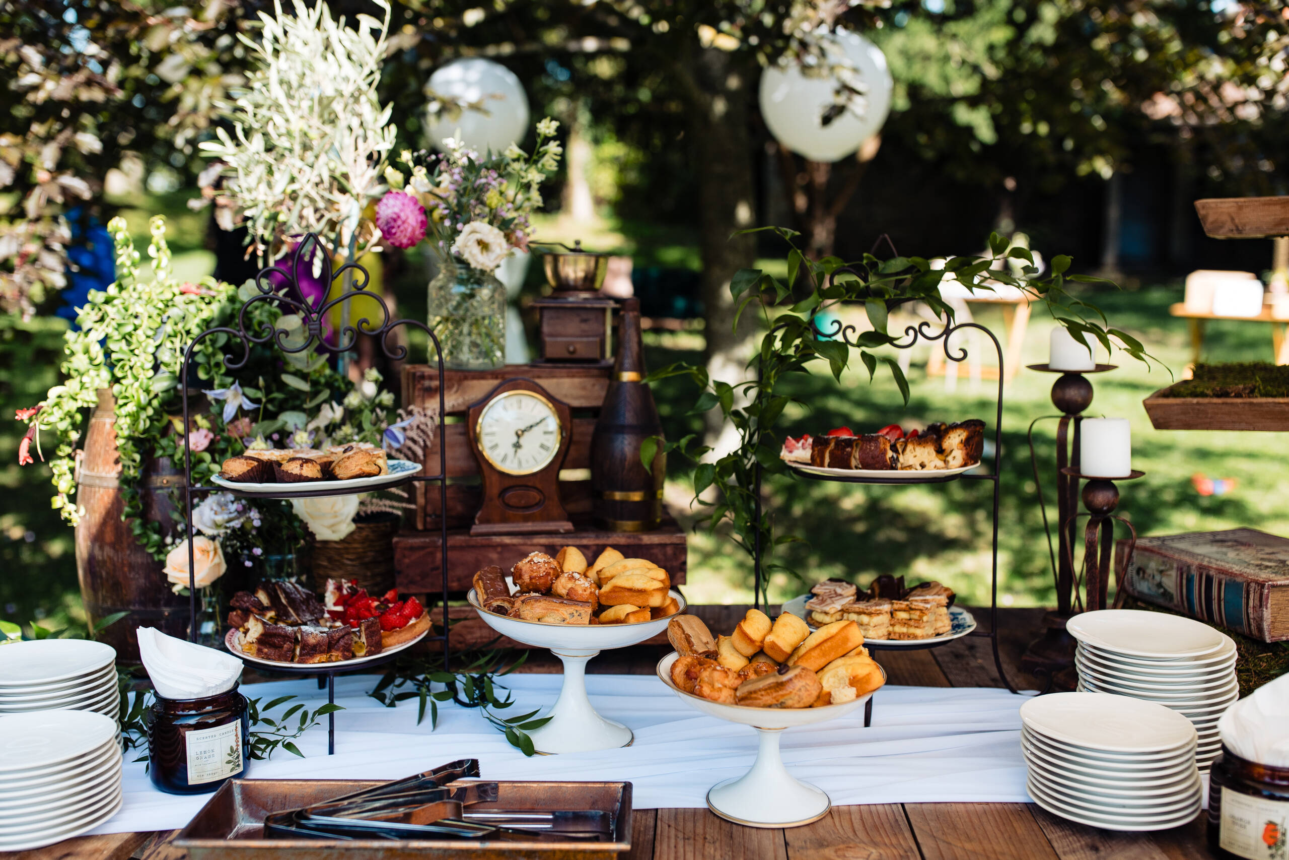 Outdoor garden party table full of pastries, flowers, and vintage decor—clock centerpiece under leafy trees.