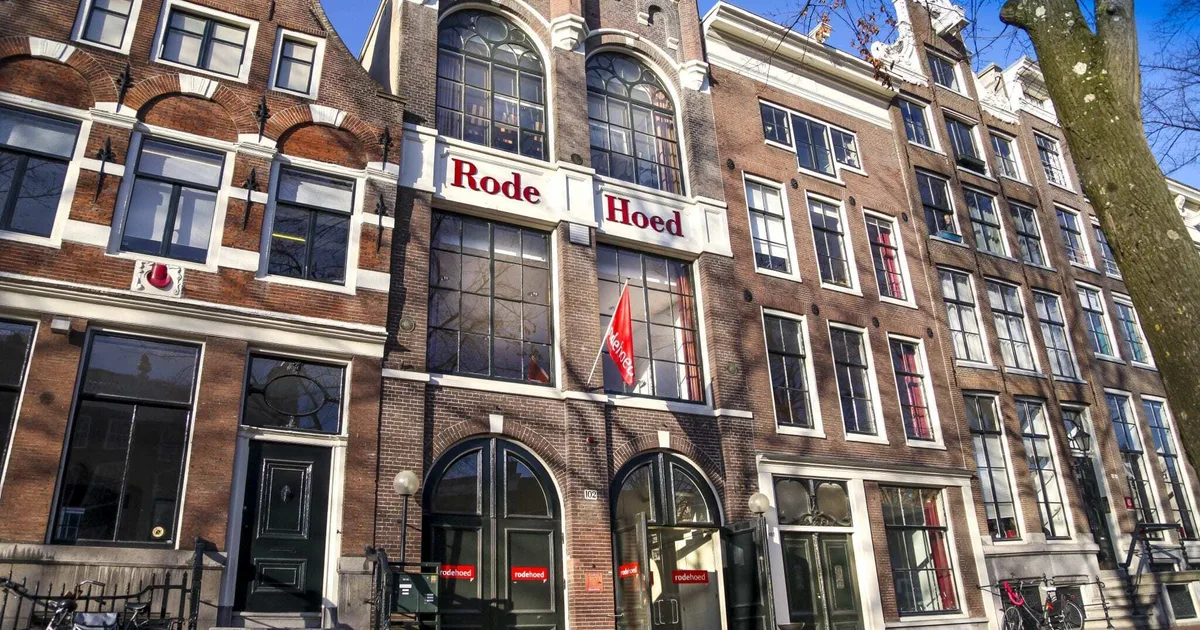 Brick canal-side building with arched windows and a prominent red 'Rode Hoed' sign above the entrance, urban Dutch street scene.