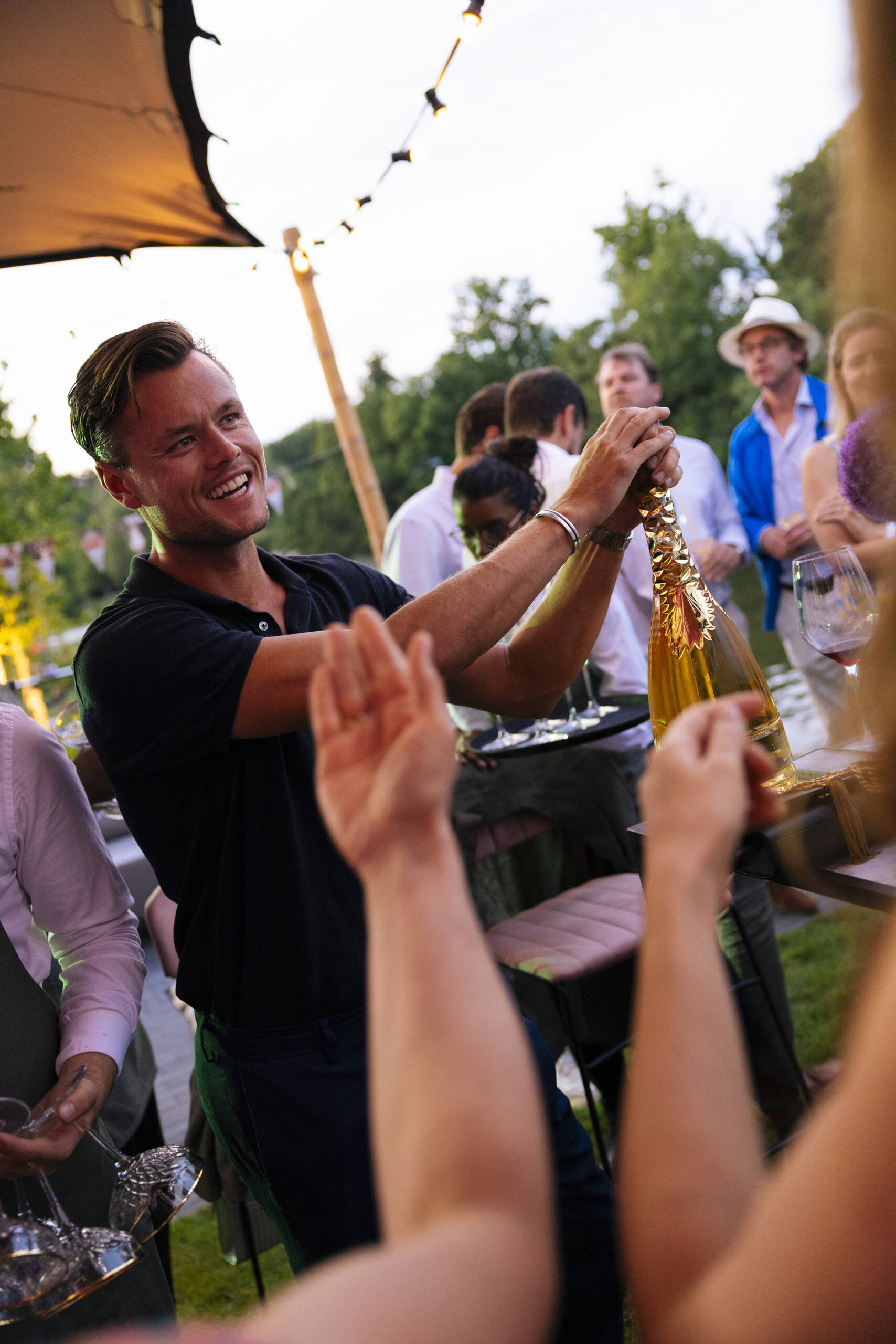 Smiling man in a black polo toasting with a champagne bottle at an outdoor party with string lights and guests in the background.