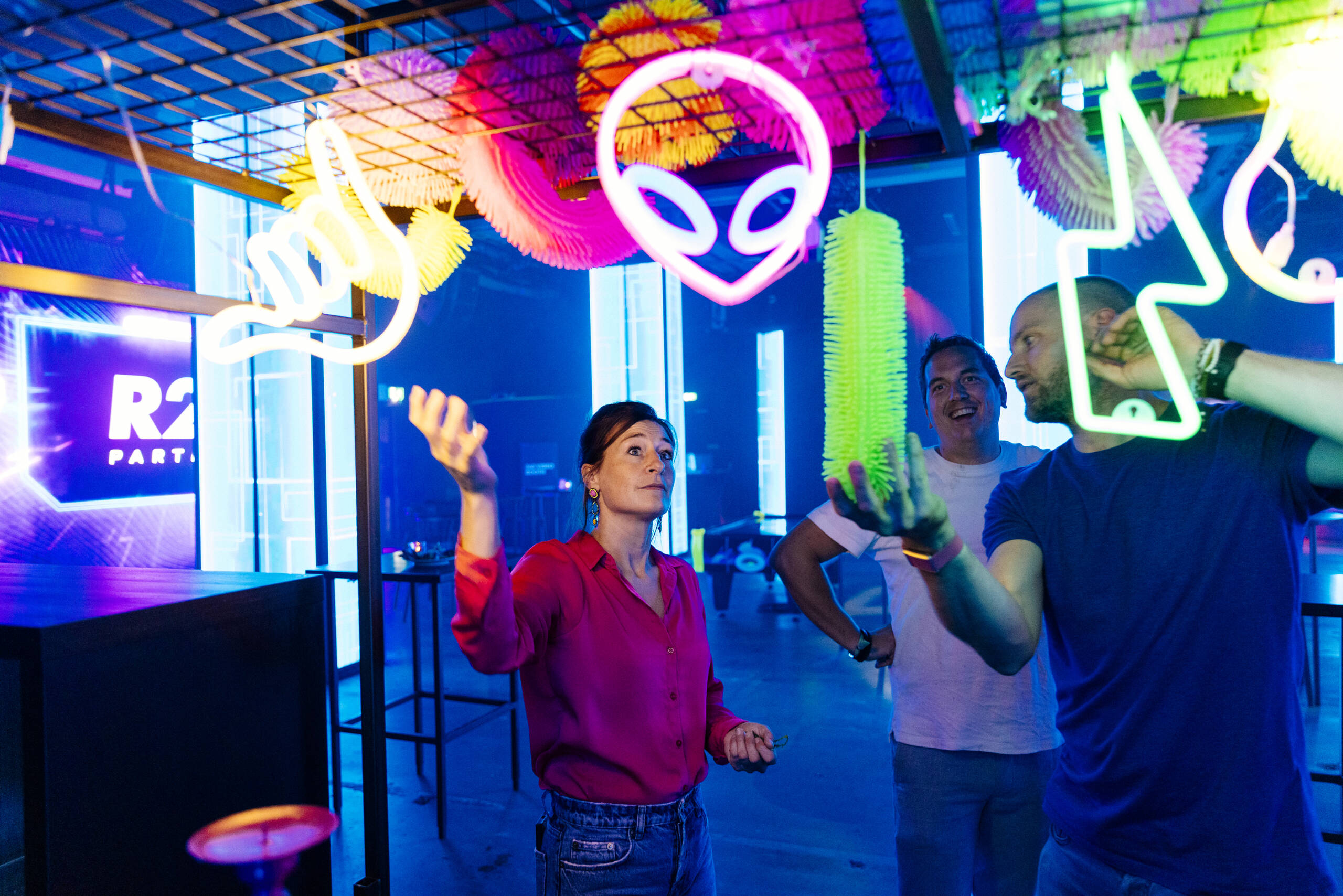 Group of friends at a neon-lit arcade, the woman in red gesturing while others smile under colorful glowing signs overhead.